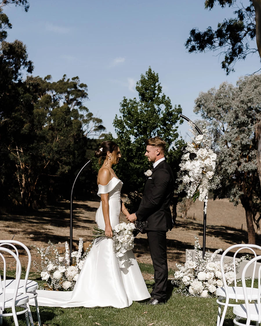 A bride and groom standing behind a black arbour with white flowers overlooking a vineyard in the Adelaide Hills