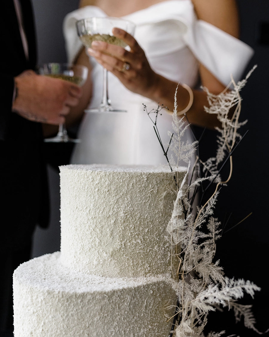 a bride and groom who are holding a glass of champagne each standing next to a cream coloured wedding cake