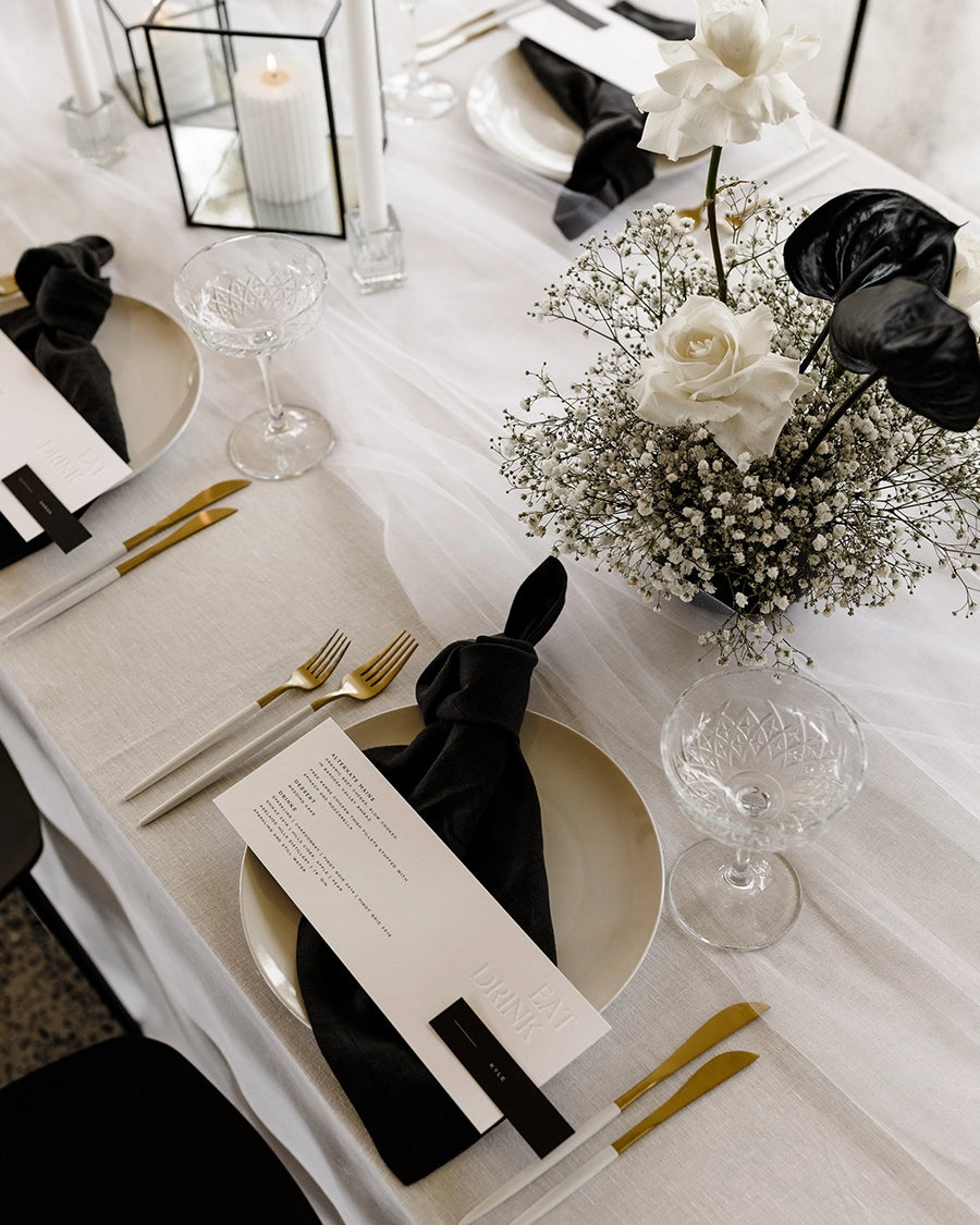 a table setting with gold cutlery, black napkin and white letterpressed menu card with a floral arrangement of babies breathe and cream roses